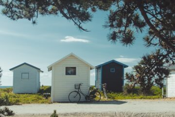 three tiny houses with a bike leaning against one; there's a tree stretching over the skyline and a few clouds in the sky