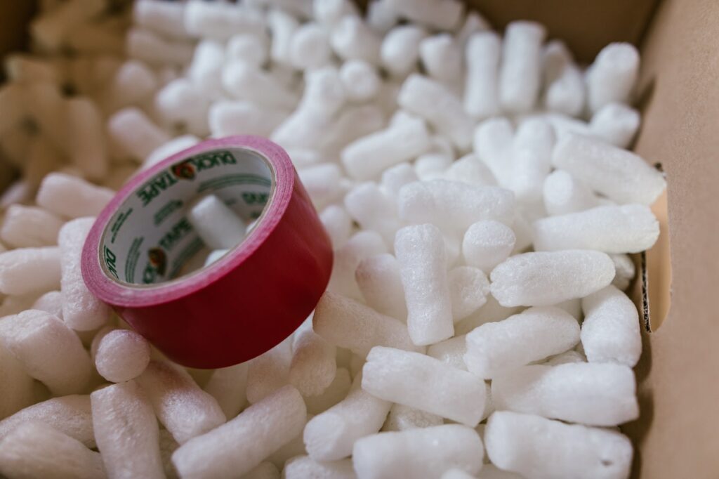 A red roll of packing tape sitting atop a pile of white packing peanutes inside a cardboard box.