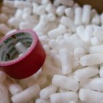 A red roll of packing tape sitting atop a pile of white packing peanutes inside a cardboard box.