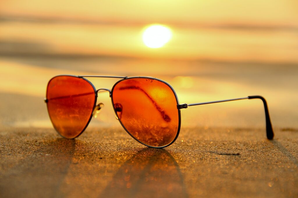 Red sunglasses sitting on the sand at a beach with the sun setting behind it.