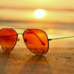 Red sunglasses sitting on the sand at a beach with the sun setting behind it.