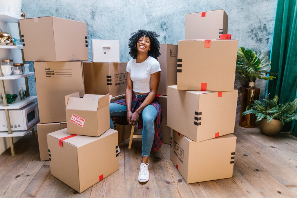 A Black woman wearing jeans and a white shirt, sitting between piles of cardboard boxes and smiling.