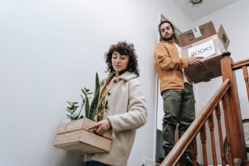 Two people walking down the stairs; one is holding a box of plants, the other a box labeled "books"