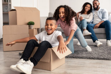 Portrait of happy black family playing and celebrating relocation, laughing daughter riding her brother in carton container, parents hugging while sitting on couch in background