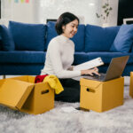 Young asian woman using laptop and taking notes while organizing clothes into boxes for storage and moving house. She is smiling and looking at the laptop screen