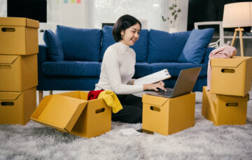 Young asian woman using laptop and taking notes while organizing clothes into boxes for storage and moving house. She is smiling and looking at the laptop screen